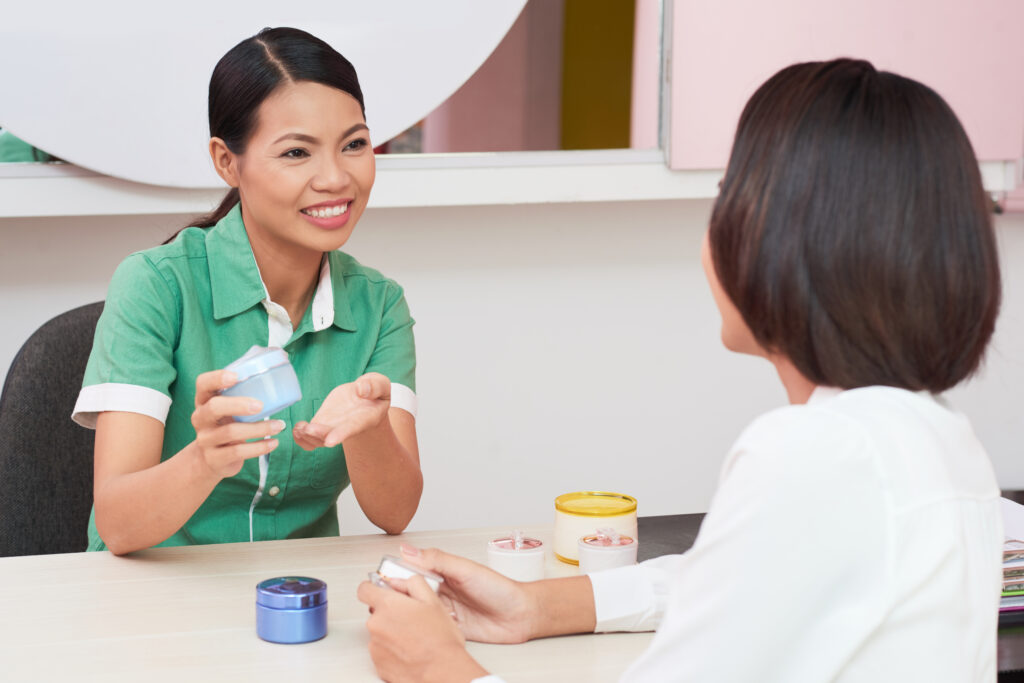 Pretty Vietnamese beautician demonstrating new cream to young woman